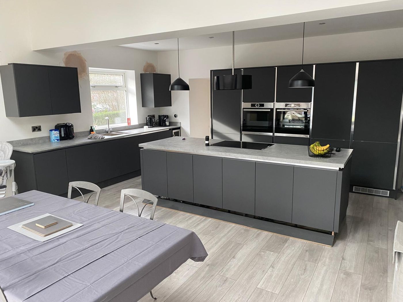 Large handleless anthracite grey kitchen with a long island, grey stone-effect worktops, tall housings with two built-in ovens and black pendant lights