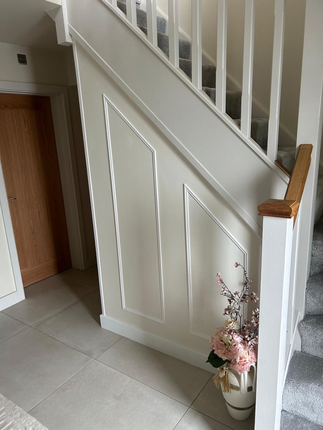 Staircase side wall finished in white panelling, white spindles and an oak handrail cap, grey tiled hallway floor